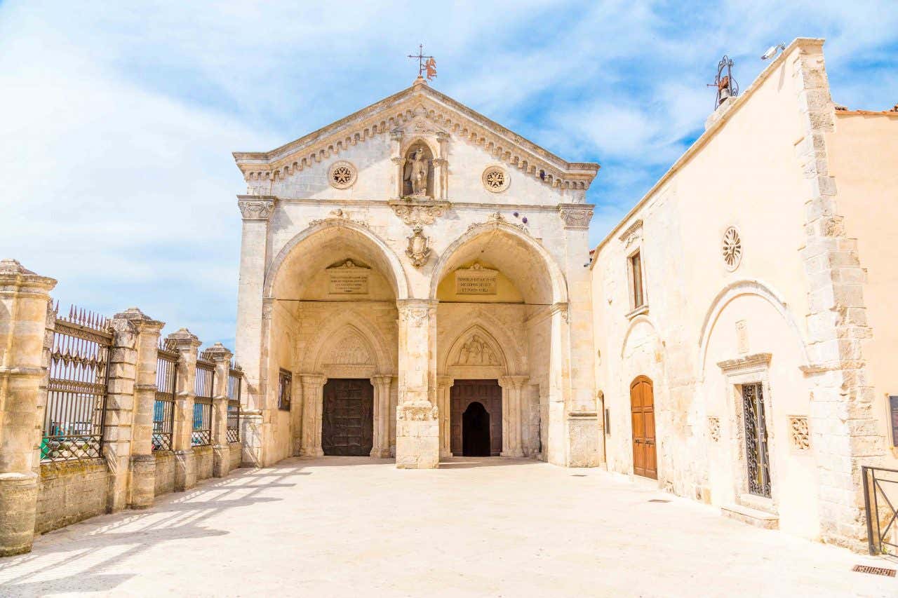 Vista frontale della facciata superiore del Santuario di San Michele Arcangelo a Monte Sant’Angelo, con un portico di due ampie arcate ogivali in pietra chiara, sormontato da un frontone triangolare decorato con archetti pensili. Al centro del frontone è incastonata una nicchia gotica contenente la statua in pietra dell’Arcangelo Michele, affiancata da due piccoli rosoni. Sulla destra si erge il campanile ottagonale