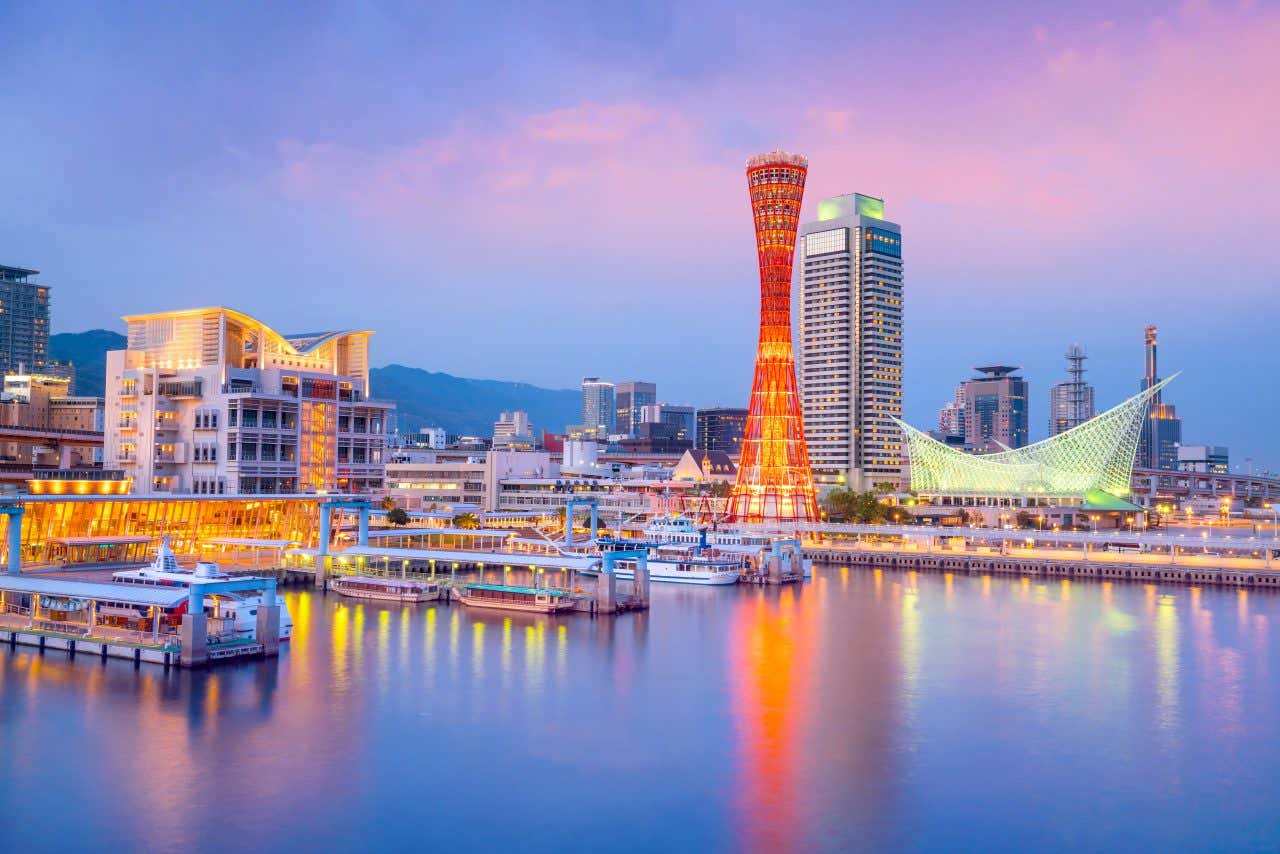 The Kobe Port Tower, a red latticework structure, stands prominently in the Kobe Harborland skyline at dusk. Modern buildings with illuminated windows are visible along the waterfront, with reflections on the water and mountains in the background.