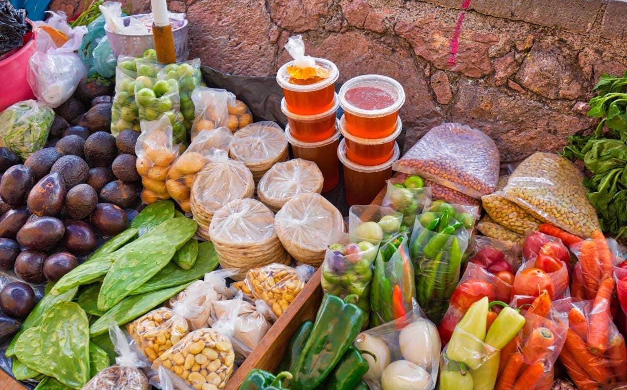 A close-up of a vibrant market stall displaying a variety of fresh produce and packaged goods. Items include avocados, nopal paddles, carrots, peppers, and stacks of tortillas.