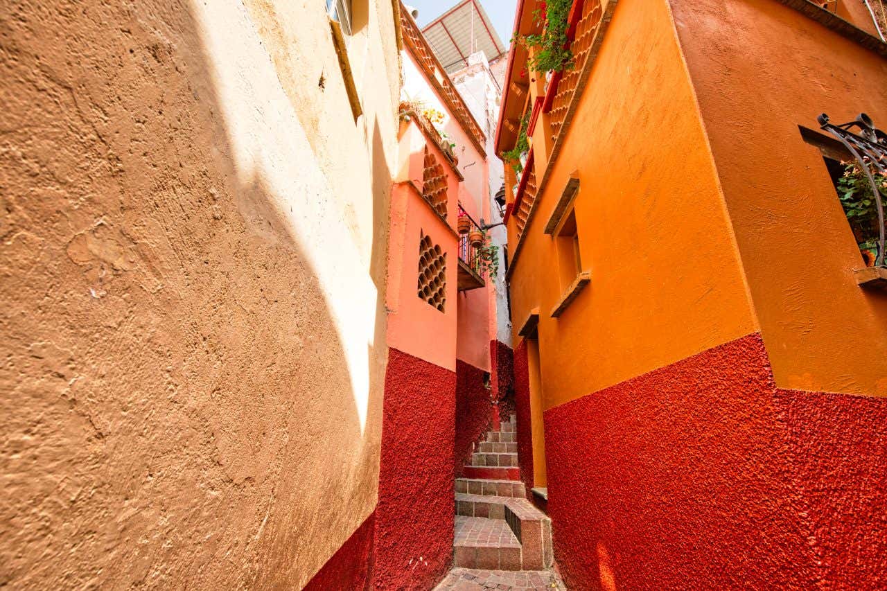 A narrow, stepped alleyway in Guanajuato, Mexico, framed by tall buildings with orange and red textured walls. Sunlight illuminates one side of the alley, while the other remains in shadow.