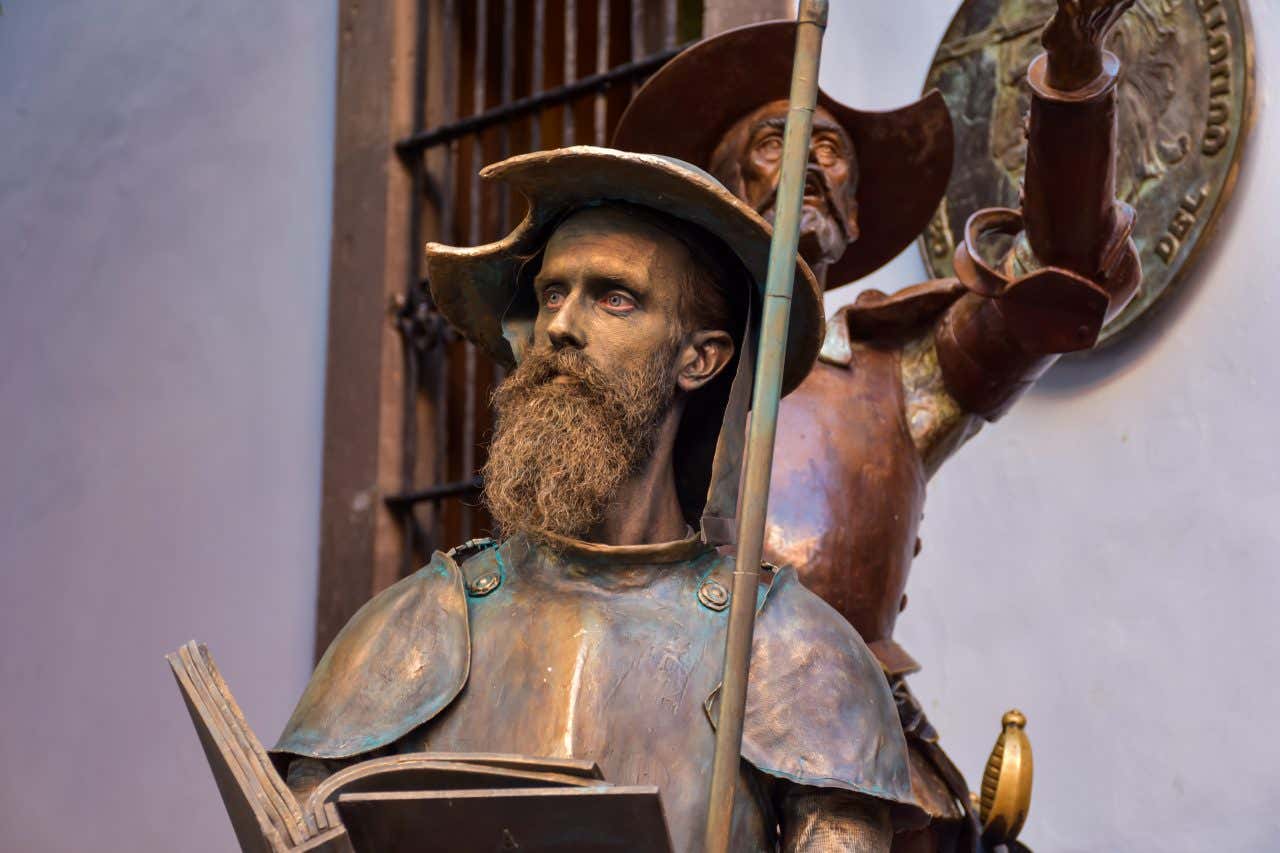 A close-up of a bronze statue depicting Don Quixote, recognizable by his helmet, beard, and a book he holds. Another statue, likely Sancho Panza, is partially visible behind him.