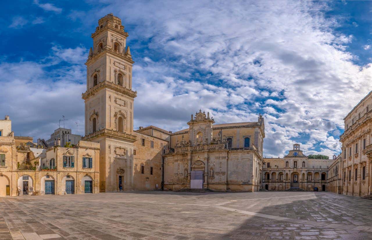 Piazza del Duomo a Lecce in una luminosa giornata di sole, con la Cattedrale barocca in primo piano, la pavimentazione chiara che riflette la luce e gli edifici storici che incorniciano la piazza