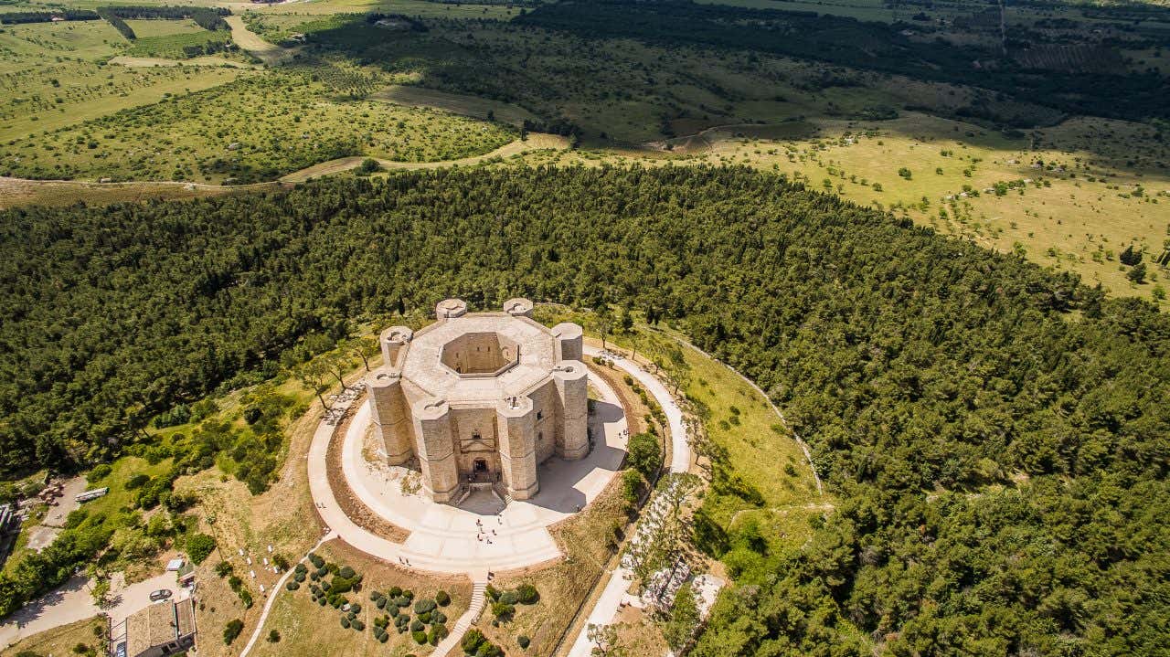 Veduta aerea della fortezza ottagonale di Castel del Monte. Si intravede chiaramente la struttura centrale ottagonale circondata da otto torri anch'esse ottagonali, disposte simmetricamente. L’edificio in pietra chiara è circondato da un percorso circolare e da terreno collinare prevalentemente arido e cespuglioso
