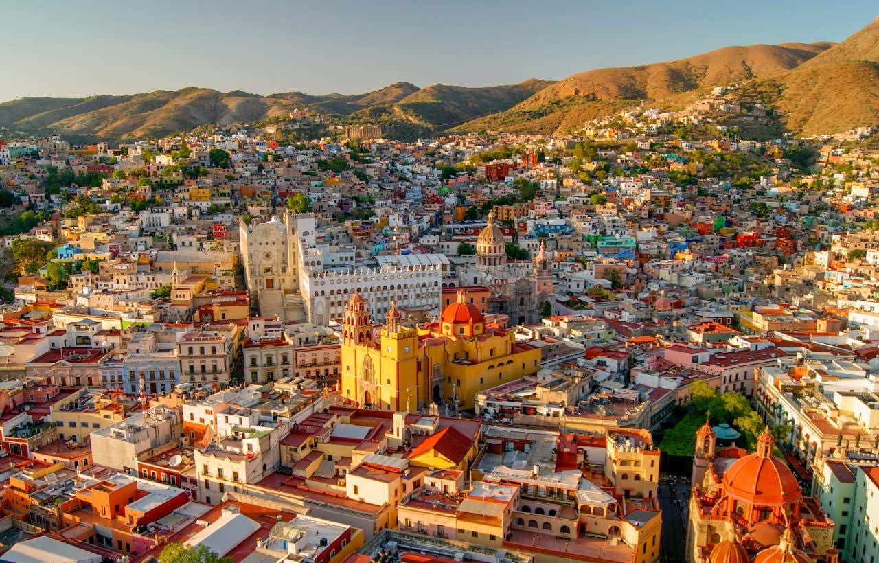 An elevated view of Guanajuato, Mexico, showing a sprawling city built into hillsides. Numerous colorful buildings with red rooftops fill the landscape, with larger white and yellow church structures visible in the foreground.