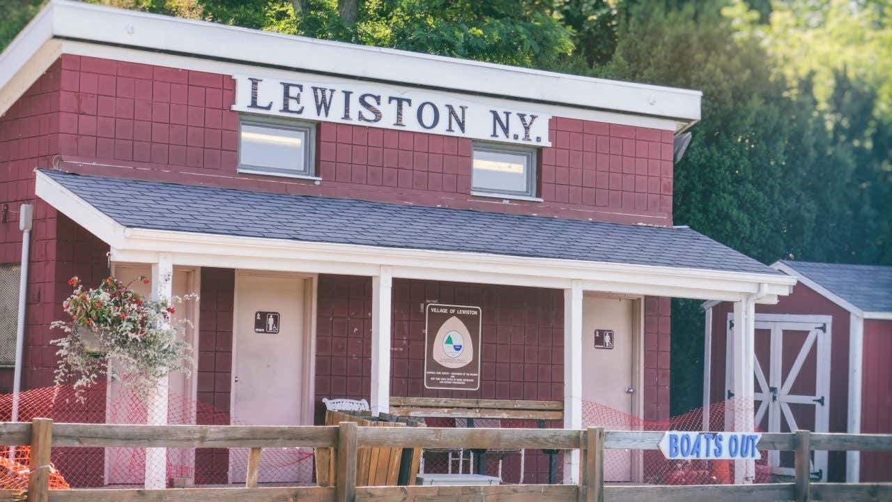 A red building with a white roof and porch, featuring a sign that reads "LEWISTON N.Y." above the windows. Two doors with restroom symbols are visible, and a wooden fence is in the foreground.