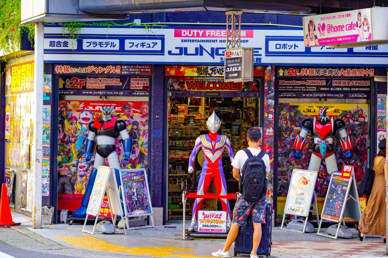 The storefront of "Jungle" a duty-free entertainment shop in Den Den Town, Osaka, Japan. The entrance is flanked by large robot and superhero statues, and a person with a backpack and luggage stands in front of the shop.