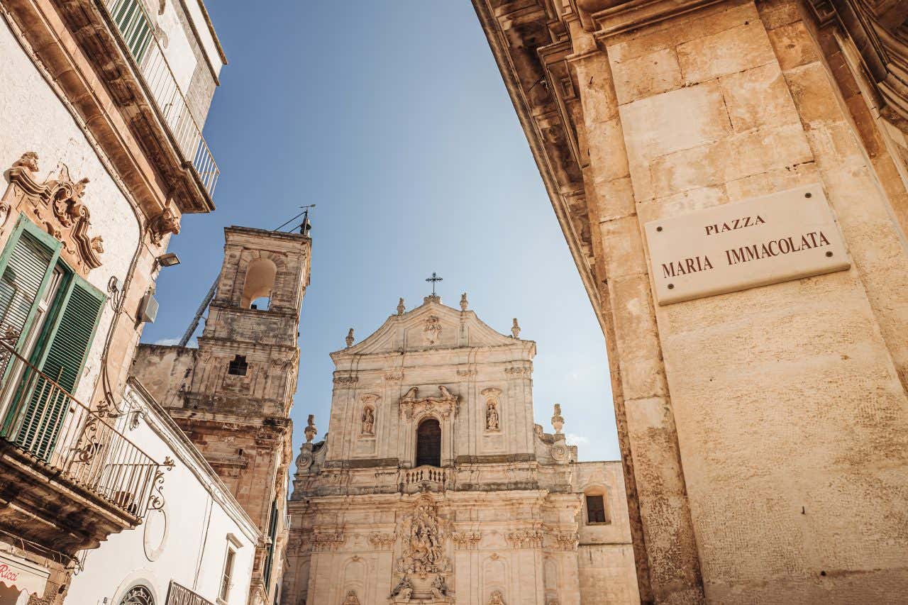 Veduta del centro storico di Martina Franca con la facciata barocca della Basilica di San Martino sullo sfondo e un'insegna in primo piano che indica 'Piazza Maria Immacolata'. L’architettura è caratterizzata da edifici in pietra chiara, balconi in ferro battuto e dettagli decorativi sotto un cielo limpido