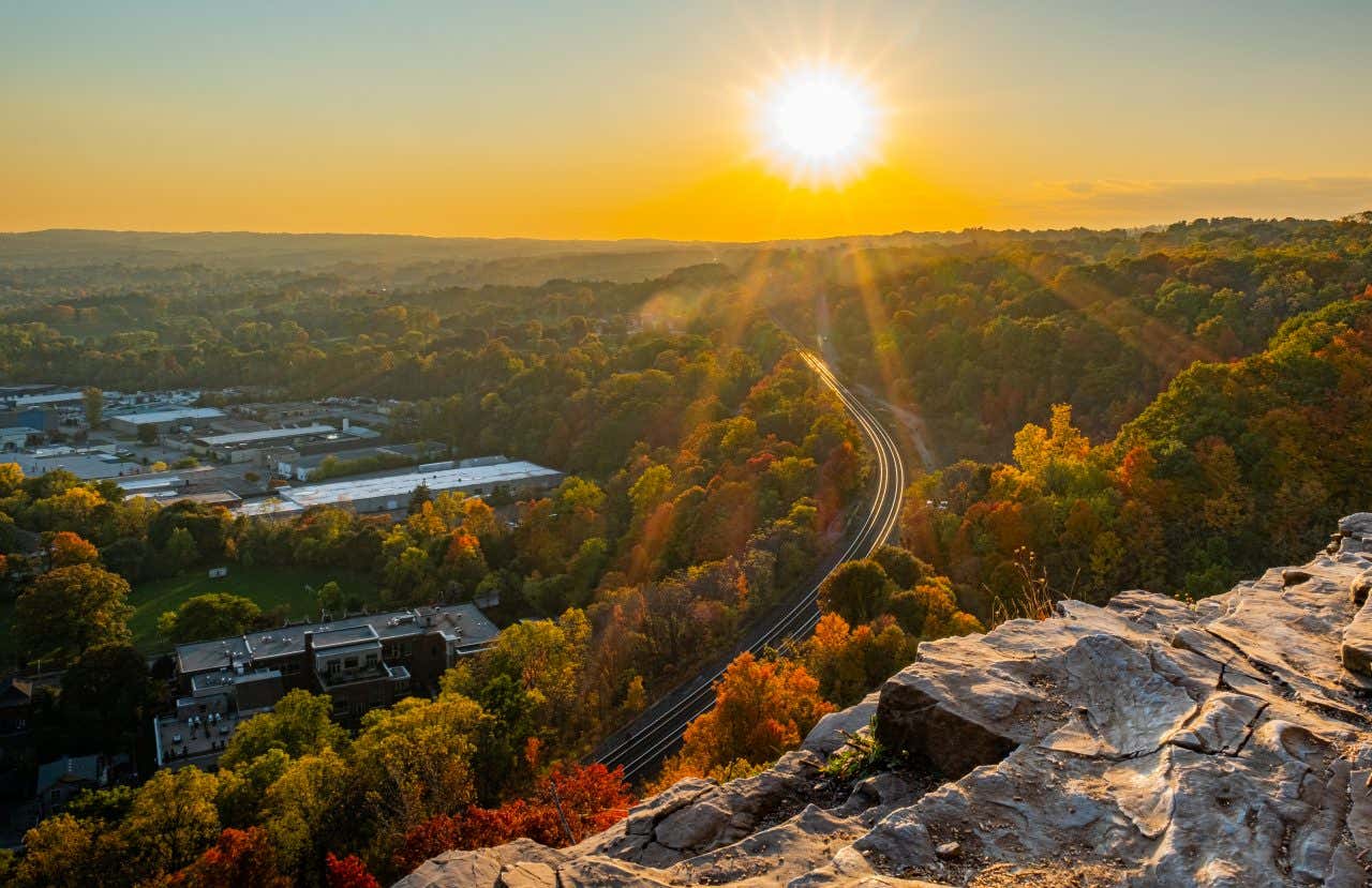 A high-angle view from Dundas Peak in Hamilton, showing a winding railway track cutting through a valley filled with trees displaying autumn foliage. The setting sun is visible in the sky, casting rays over the landscape, with urban areas in the distance.