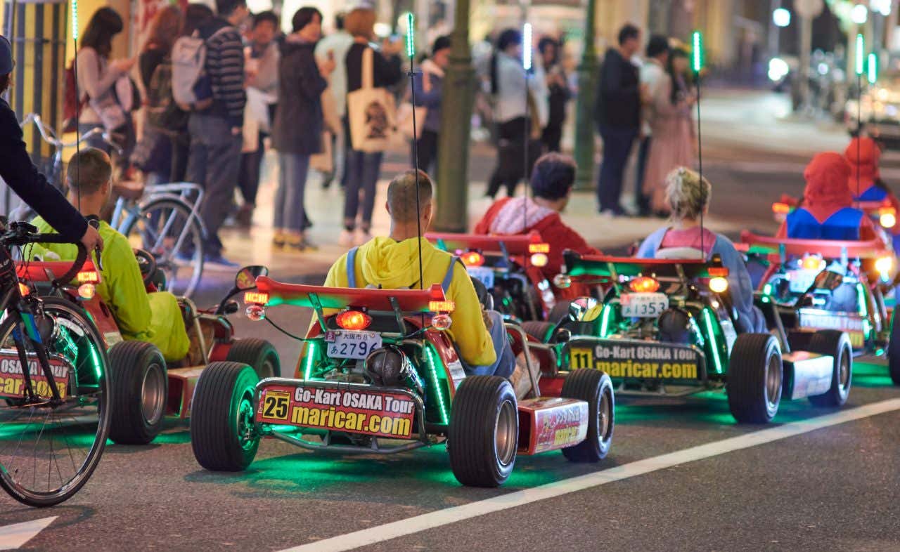 People in go-karts, some dressed in character costumes, are driving on a city street at night. The go-karts have green lights, and pedestrians are visible on the sidewalk in the background.