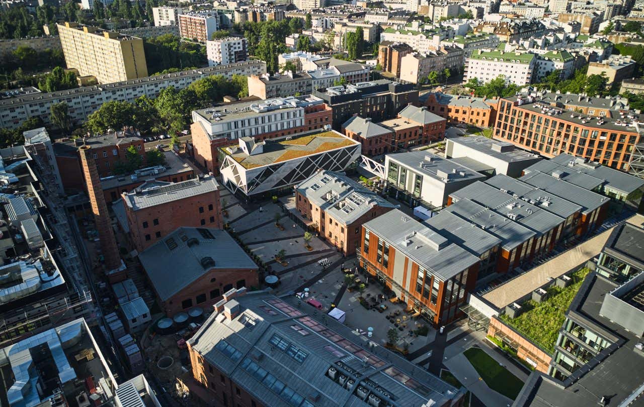 Vista aerea della Piazza Koneser nel quartiere Praga di Varsavia, con edifici in mattoni rossi ristrutturati in stile industriale, moderni complessi commerciali e spazi pubblici organizzati attorno a una piazza centrale pedonale. L’area combina architettura storica e contemporanea, mostrando un vivace centro culturale e commerciale integrato nel tessuto urbano della città