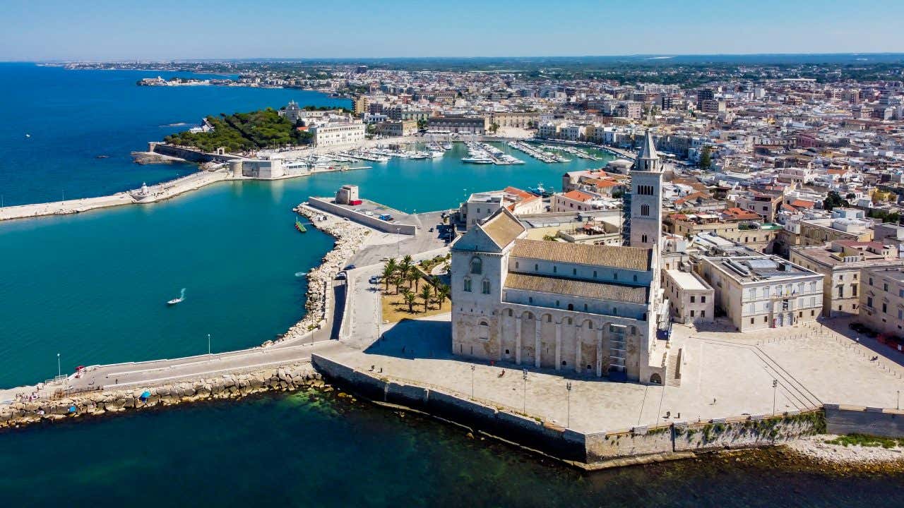 Veduta aerea della Cattedrale di Trani vista dal mare in una giornata di sole: la chiesa romanica in calcare chiaro domina il porto, con la sua pianta a tre navate, la facciata sollevata e la grande torre campanaria allineata al lato, mentre la città si estende sullo sfondo tra il porto e le banchine affollate di barche
