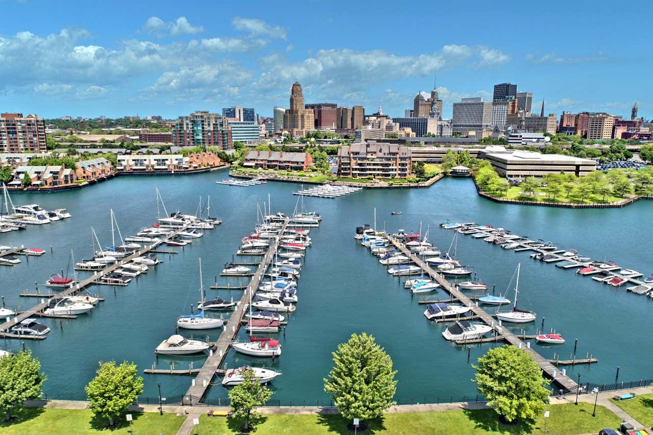 An aerial view of a marina in Buffalo, New York, filled with numerous boats docked in the blue water. Buildings and urban development line the waterfront, with the city skyline visible in the distance under a partly cloudy sky.