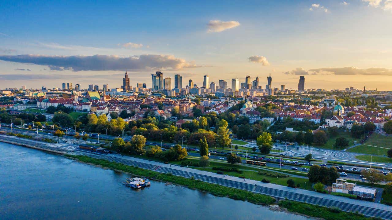 Panorama della città di Varsavia al tramonto, con il fiume Vistola in primo piano, alberi e strade trafficate in basso, e il moderno skyline con grattacieli e edifici storici sullo sfondo