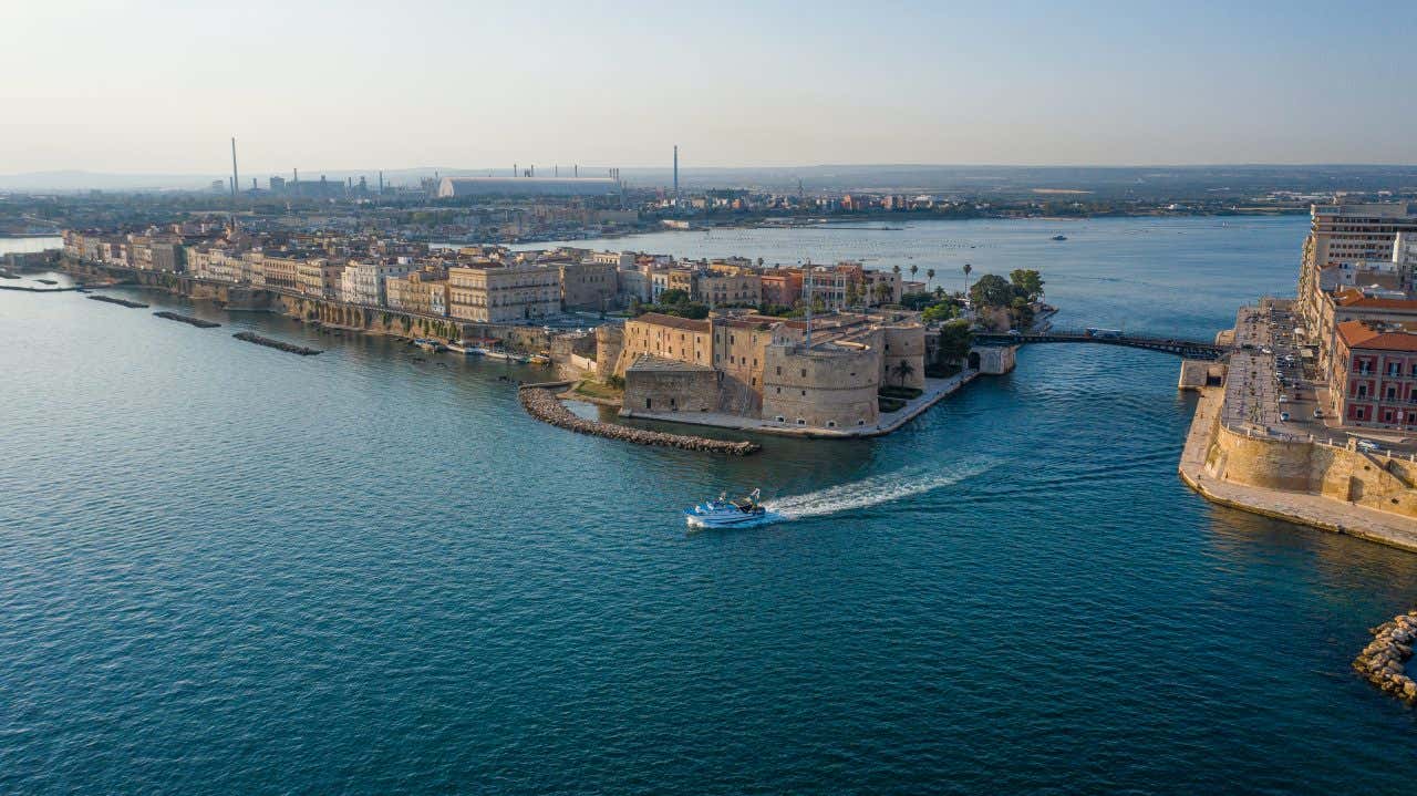 Veduta aerea di Taranto con il ponte girevole che collega l’isola del centro storico alla città nuova, sospeso sopra il Canale Navigabile. Sotto il ponte c'è la scia di una barca appena passata, con edifici storici e moderni visibili su entrambi i lati