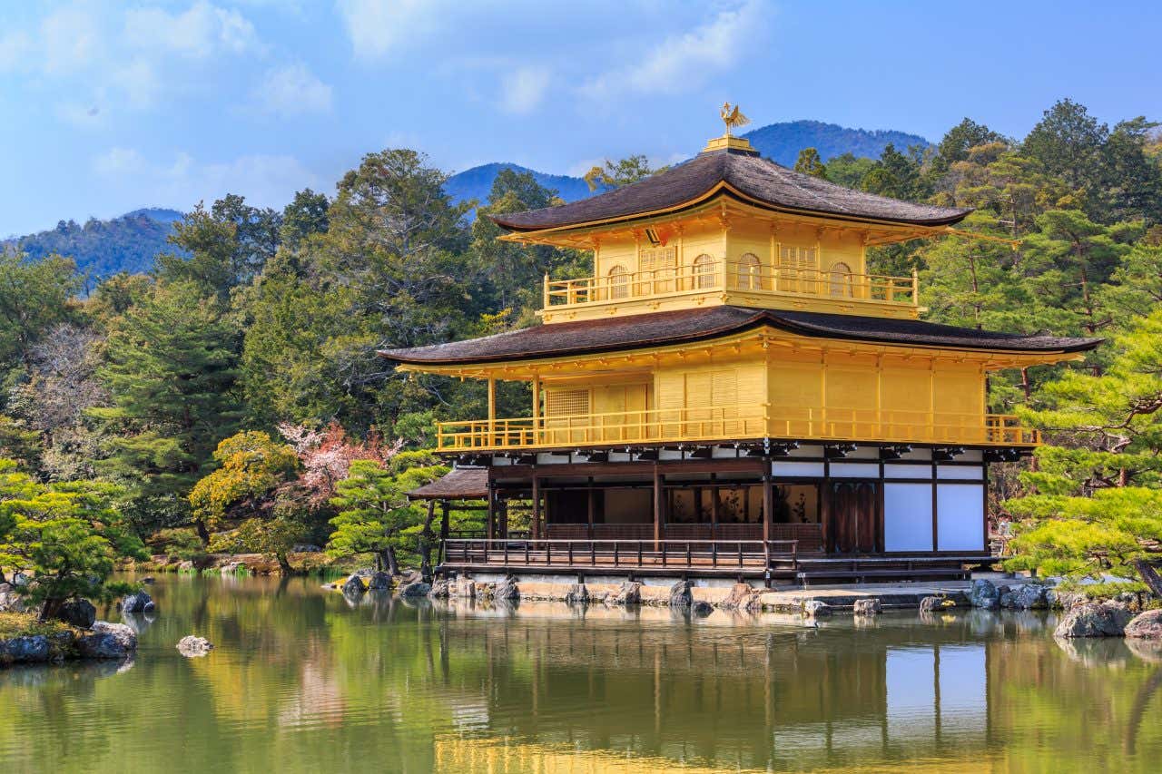 Kinkaku-ji, the Golden Pavilion temple, stands on the edge of a calm pond, with its golden exterior reflecting on the water. The temple is surrounded by green trees and foliage, and mountains are visible in the background under a blue sky.