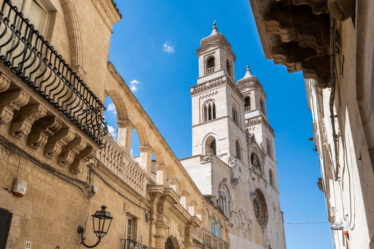 Veduta dal basso della Cattedrale di Altamura, con le sue imponenti torri campanarie gemelle in stile romanico pugliese che si ergono contro un cielo azzurro limpido; la scena è incorniciata da edifici in pietra chiara, archi medievali e balconi in ferro battuto tipici del centro storico