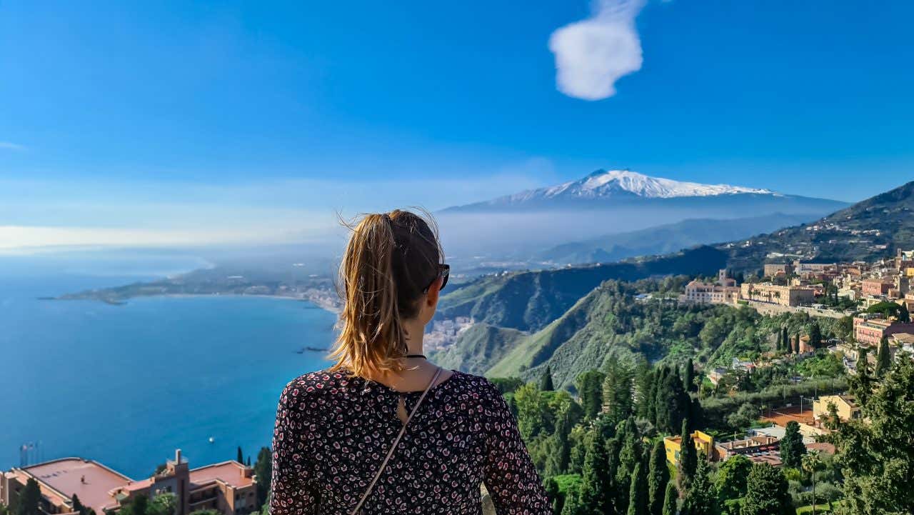A woman with her back turned admiring the views of the town of Taormina with the sea and Mount Etna in the distance.