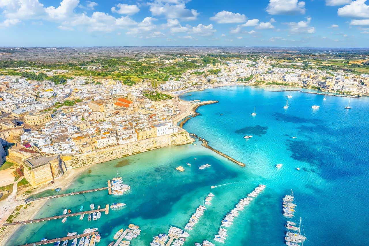 Veduta aerea del Golfo di Otranto in una luminosa giornata estiva, con il porto in primo piano e il centro storico che si sviluppa alle sue spalle, con le mura medievali fortificate affacciate sul mare azzurro