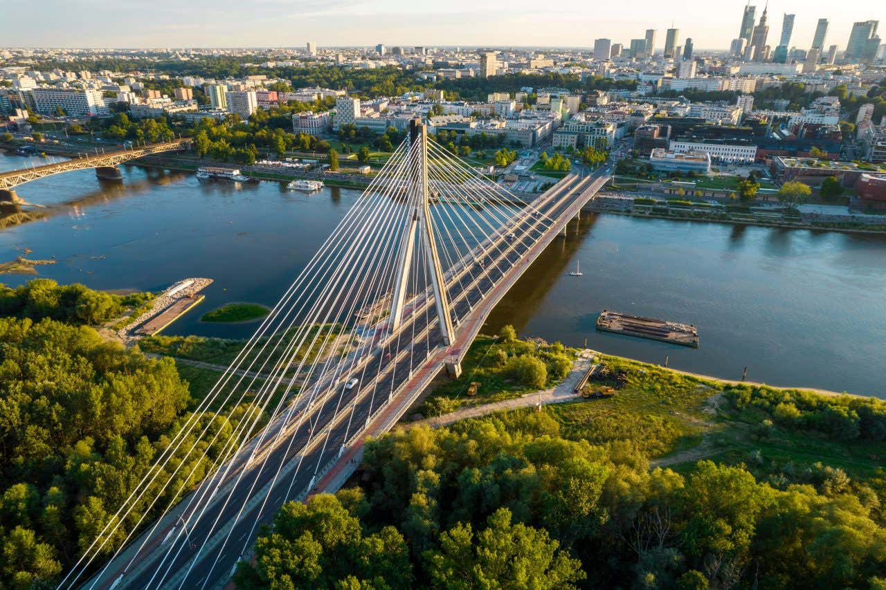 Vista al tramonto di un moderno ponte bianco su un fiume, che collega due sponde della città di Varsavia, una con molti edifici e l'altra più verde, senza palazzi. Sul fiume navigano alcuni battelli, mentre sul ponte ci sono due corsie attraversate da alcune automobili