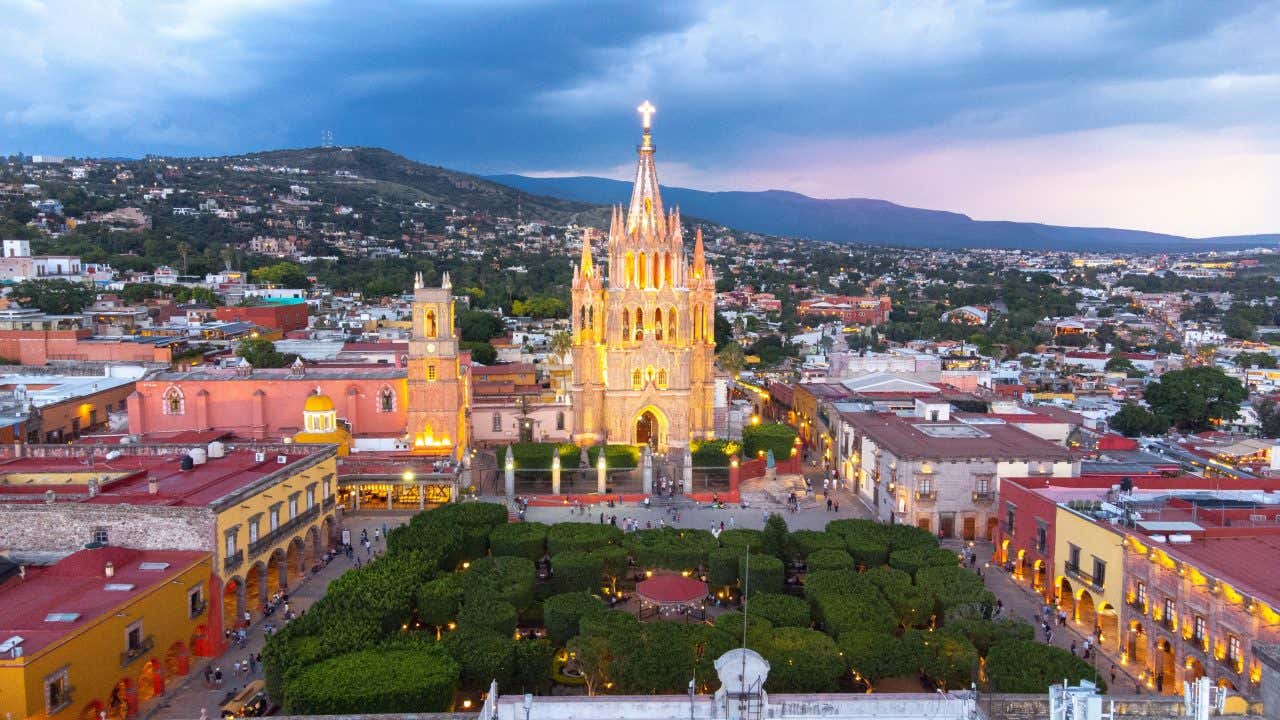An aerial view of San Miguel de Allende, Mexico, at dusk, centered on the illuminated Parroquia de San Miguel Arcángel church with its tall, spired facade. A town square with manicured green trees is in front of the church, surrounded by other city buildings and distant mountains.