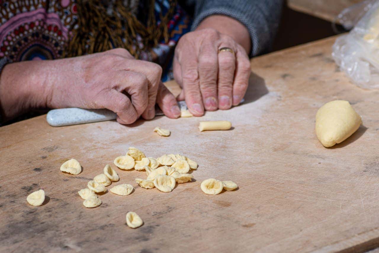 Mani di un'anziana signora che lavorano un panetto di pasta fresca su una tavola di legno con l'aiuto di un coltello per dare forma alle orecchiette