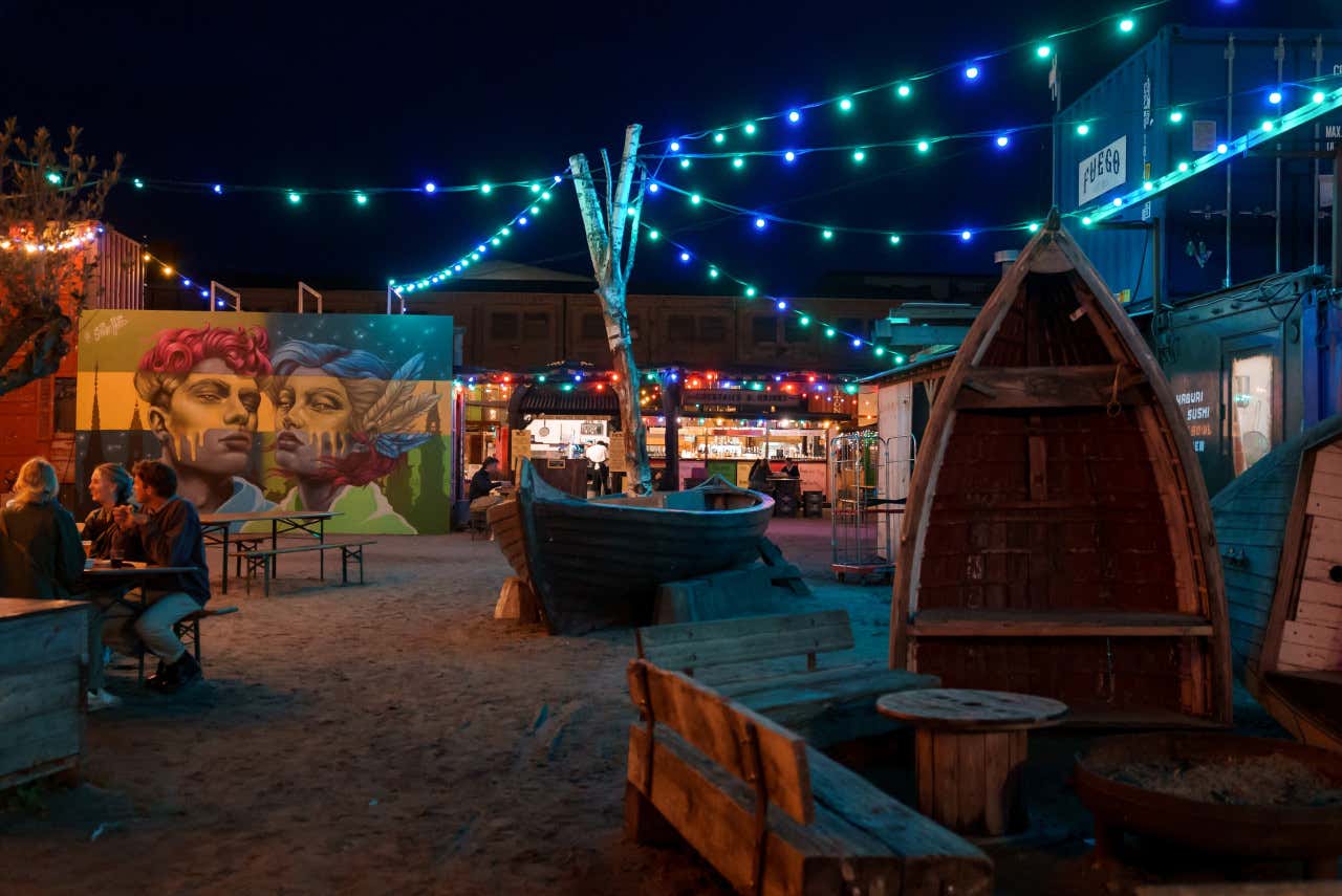 Decoração de um bar feita com barcos, mesas e cadeiras de madeira, um mural e barracas de street food ao fundo à noite em cReffen