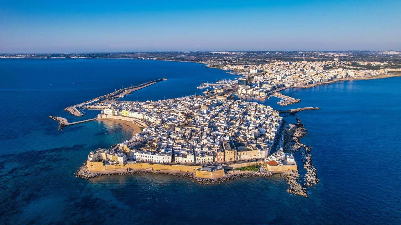 Veduta aerea del centro storico di Gallipoli ripreso dal mare, con l’antica città fortificata che si estende su un’isola collegata alla terraferma, circondata da acque cristalline. Le case bianche si affacciano sulla costa, mentre le mura e le fortificazioni seguono il profilo dell’isola