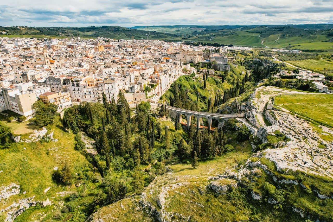 Veduta panoramica di Gravina in Puglia, con il caratteristico ponte acquedotto Madonna della Stella che attraversa una profonda gola verdeggiante, circondata da alberi e rocce. Sullo sfondo, il centro storico della città si estende con edifici in pietra chiara, mentre le colline verdi si allungano verso l’orizzonte sotto un cielo nuvoloso