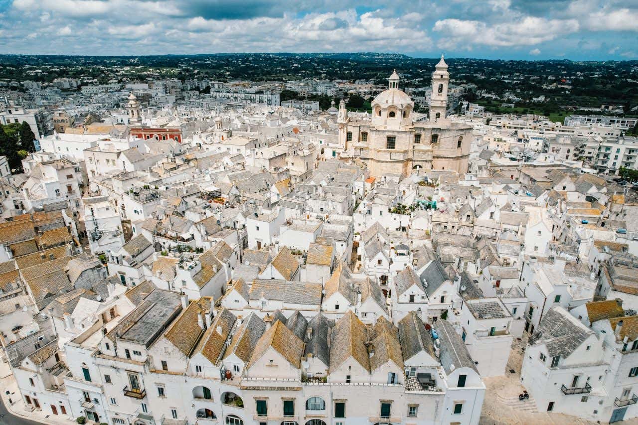 Il centro storico di Locorotondo visto dall'alto, fatto di palazzi disposti a forma circolare. Al centro del conglomerato urbano si erge la cupola e il campanile di una cattedrale, il cielo è ricoperto di nuvole bianche