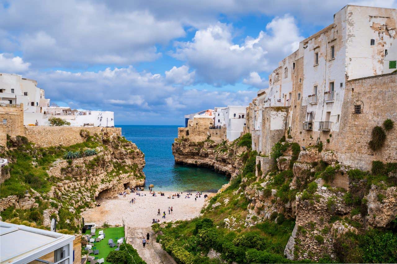 Una spiaggia piccola, chiusa ai due lati da scogliere alte sormontate dai palazzi del centro storico di Polignano a Mare. La giornata è serena, nel cielo ci sono alcune nuvole e in spiaggia pochi turisti