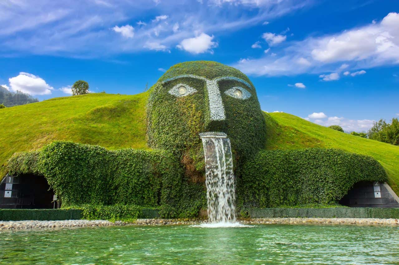 La facciata verde a forma di testa umana del museo Swarovski Kristallwelten a Wattens, Austria, con occhi luminosi e una cascata che fuoriesce dalla bocca, immersa in un paesaggio collinare sotto un cielo azzurro con nuvole sparse