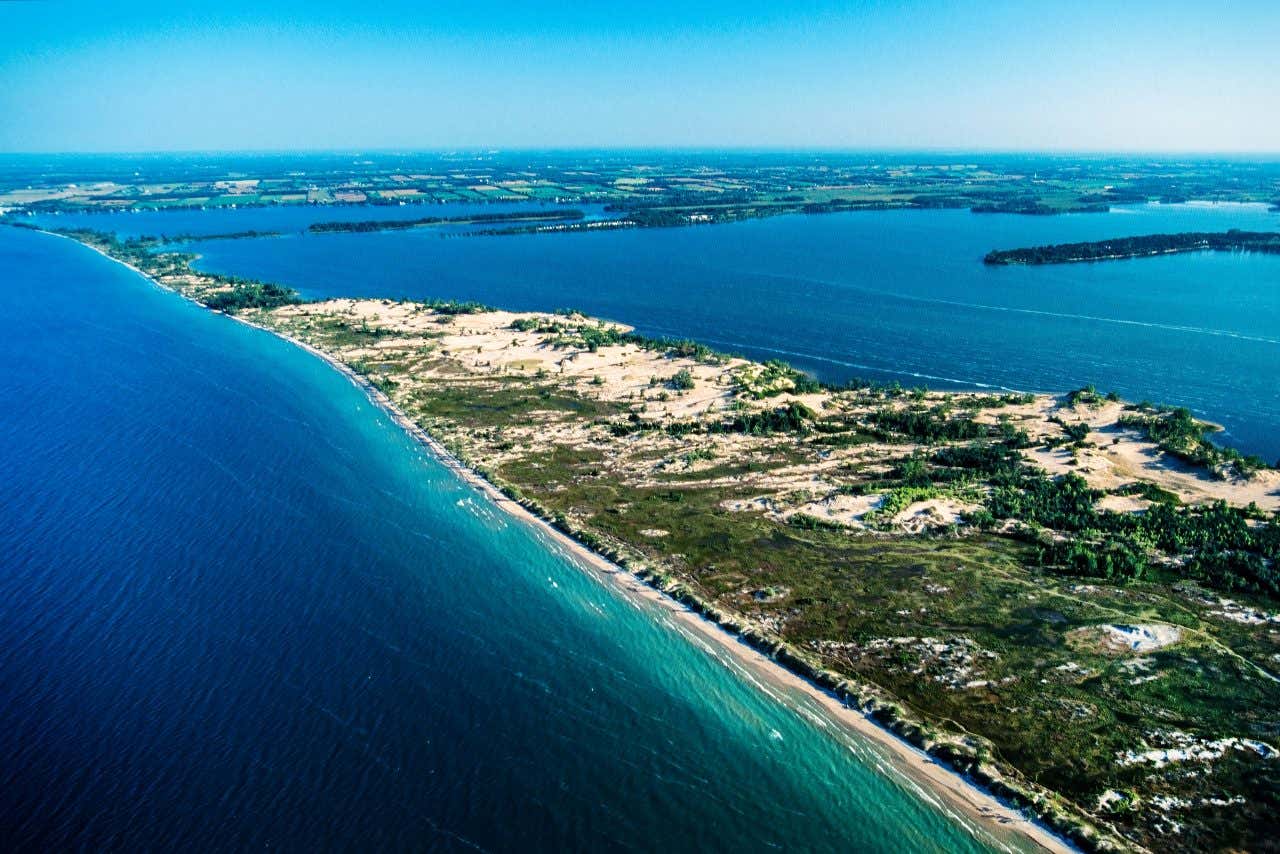 An aerial view of Sandbanks Provincial Park, showing a long strip of sandy dunes and green vegetation separating a large body of deep blue water (lake or bay) from the lighter blue water of a major lake. Farmland is visible on the distant horizon.