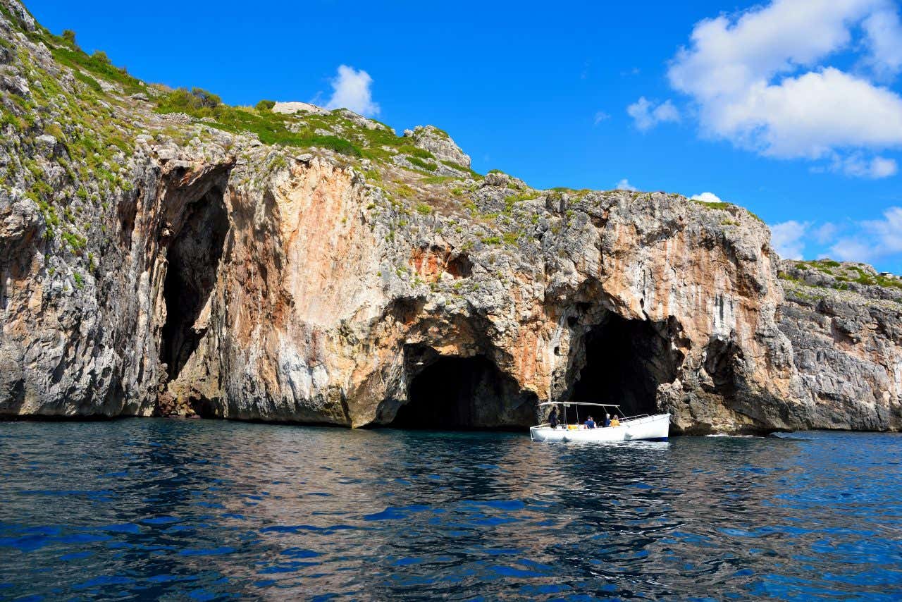 Le Grotte delle Tre Porte a Santa Maria di Leuca, con le tre maestose arcate scavate nella falesia calcarea che si aprono sul mare cristallino; una piccola barchetta turistica si avvicina dal lato destro dell'immagine, puntando verso l’ingresso centrale