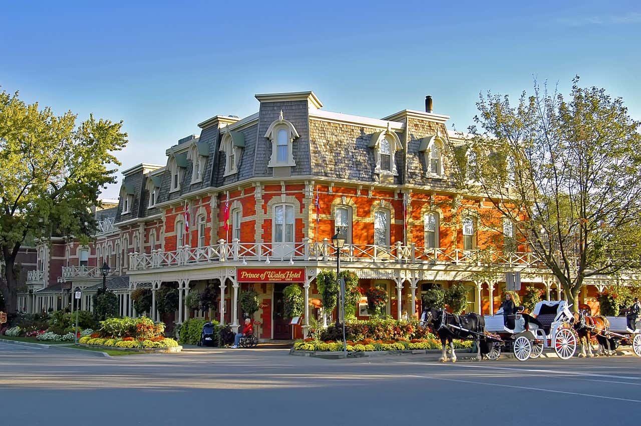 The Prince of Wales Hotel in Niagara-on-the-Lake, a red brick building with a grey roof and white trim. A horse-drawn carriage is parked in front of the hotel, and trees are visible on either side under a clear sky.