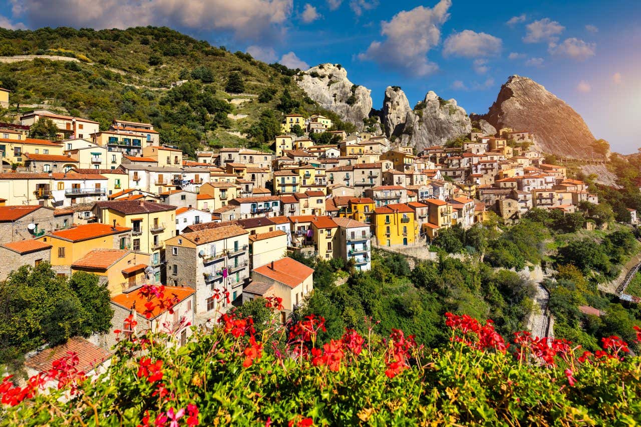 The town of Castlemezzano with houses built into the mountainside, mountains in the distance, and red flowers at the foreground.