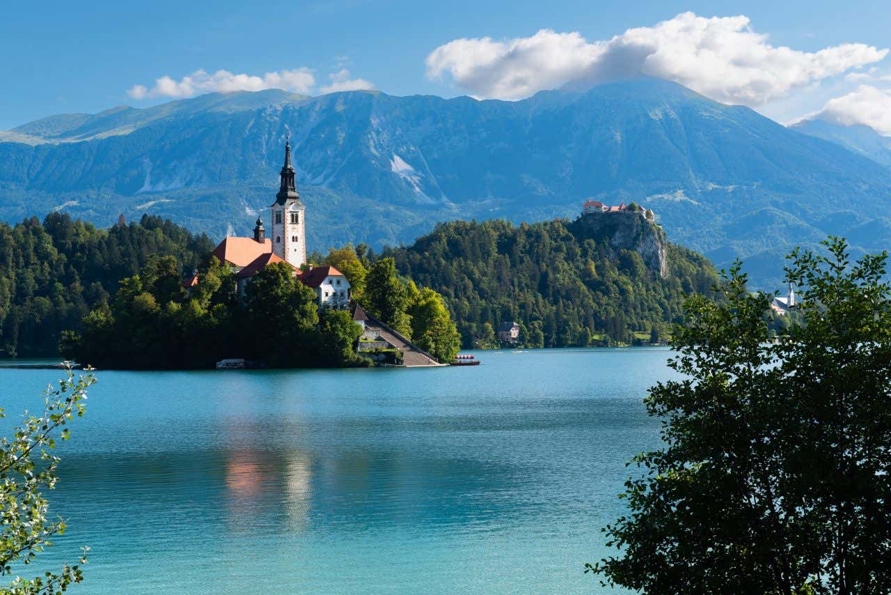 Lake Bled in Slovenia, with Bled Island and its church in the center of the turquoise water. Bled Castle is visible on a cliff overlooking the lake in the background. Mountains rise behind the lake under a partly cloudy sky.