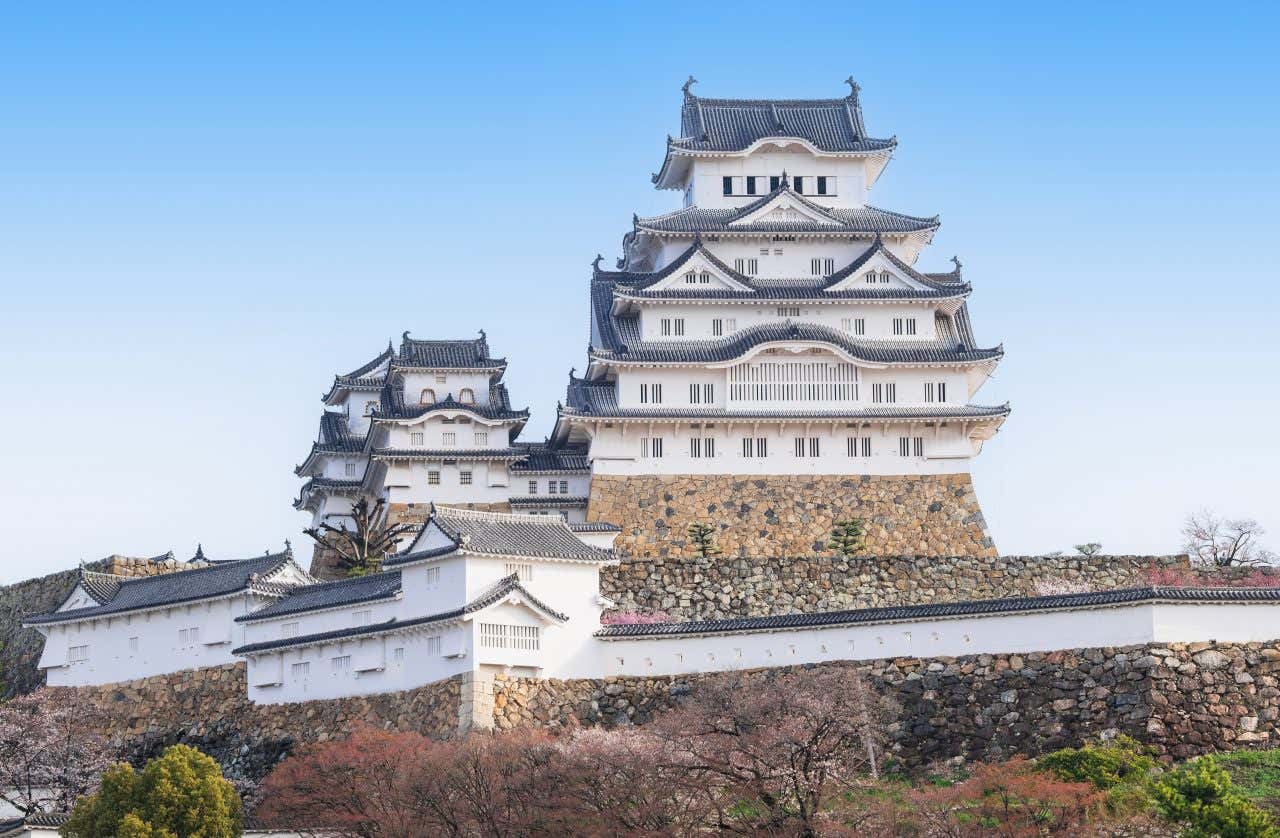 Himeji Castle, a large white Japanese castle with multiple tiers and dark grey roofs, stands atop a stone foundation. The castle is surrounded by some trees, under a clear blue sky.
