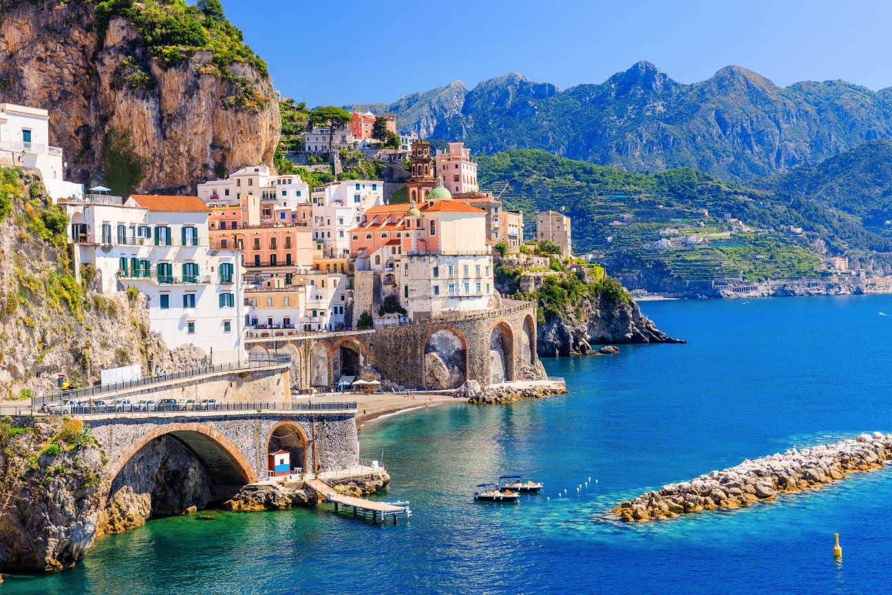 A coastline with buildings perched on a cliff next to turquoise waters, with mountains in the distance, under a clear blue sky.