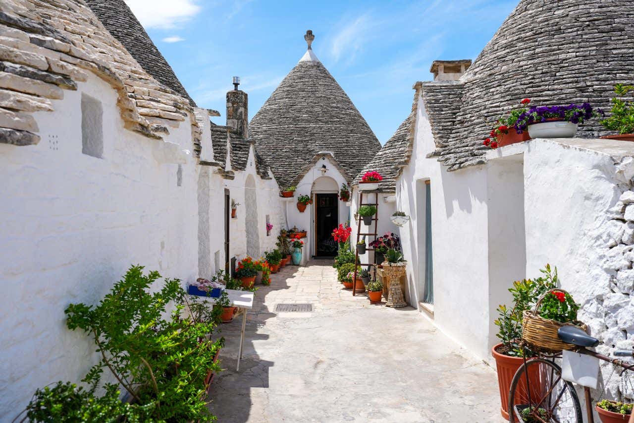 Stradina pittoresca di Alberobello fiancheggiata dai tradizionali trulli, case bianche con tetti conici in pietra. Le abitazioni sono decorate con fiori colorati in vasi e balconi, e una bicicletta con cestino fiorito è appoggiata a un muro. Il cielo è sereno con qualche nuvola