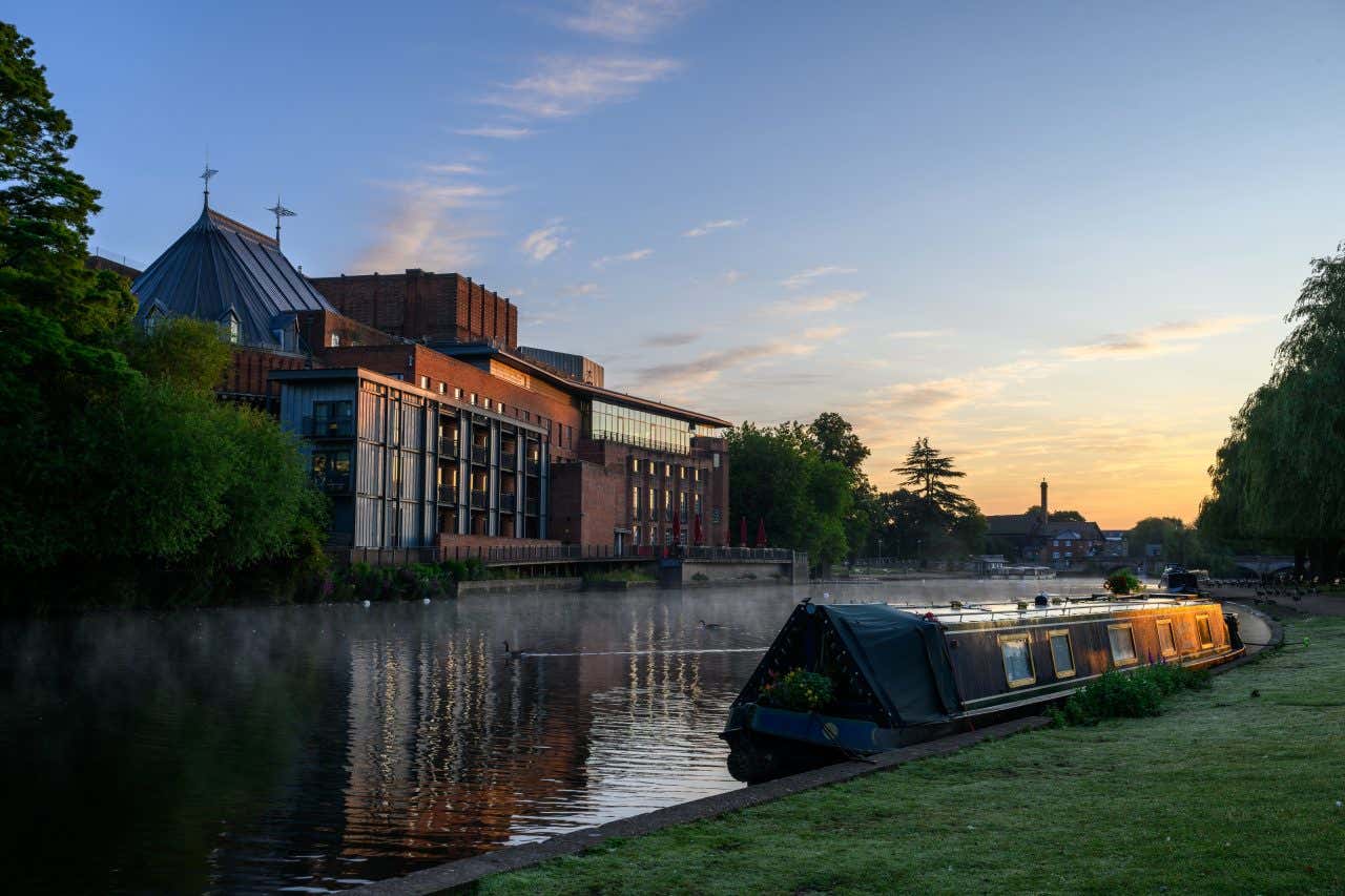 The Royal Shakespeare Theatre building, a modern brick structure with large windows, stands beside a misty river at dawn or dusk. A narrowboat is docked along the riverbank in the foreground, and trees line the far bank.