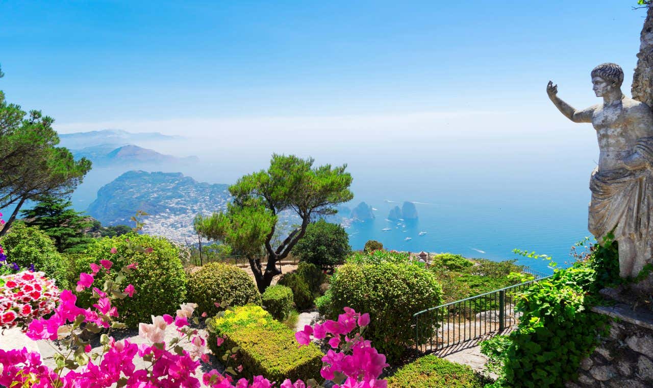 The Gardens of Augustus in Capri with a statue of Emperor Augustus and flowers with a city and large rock formations in the distance.