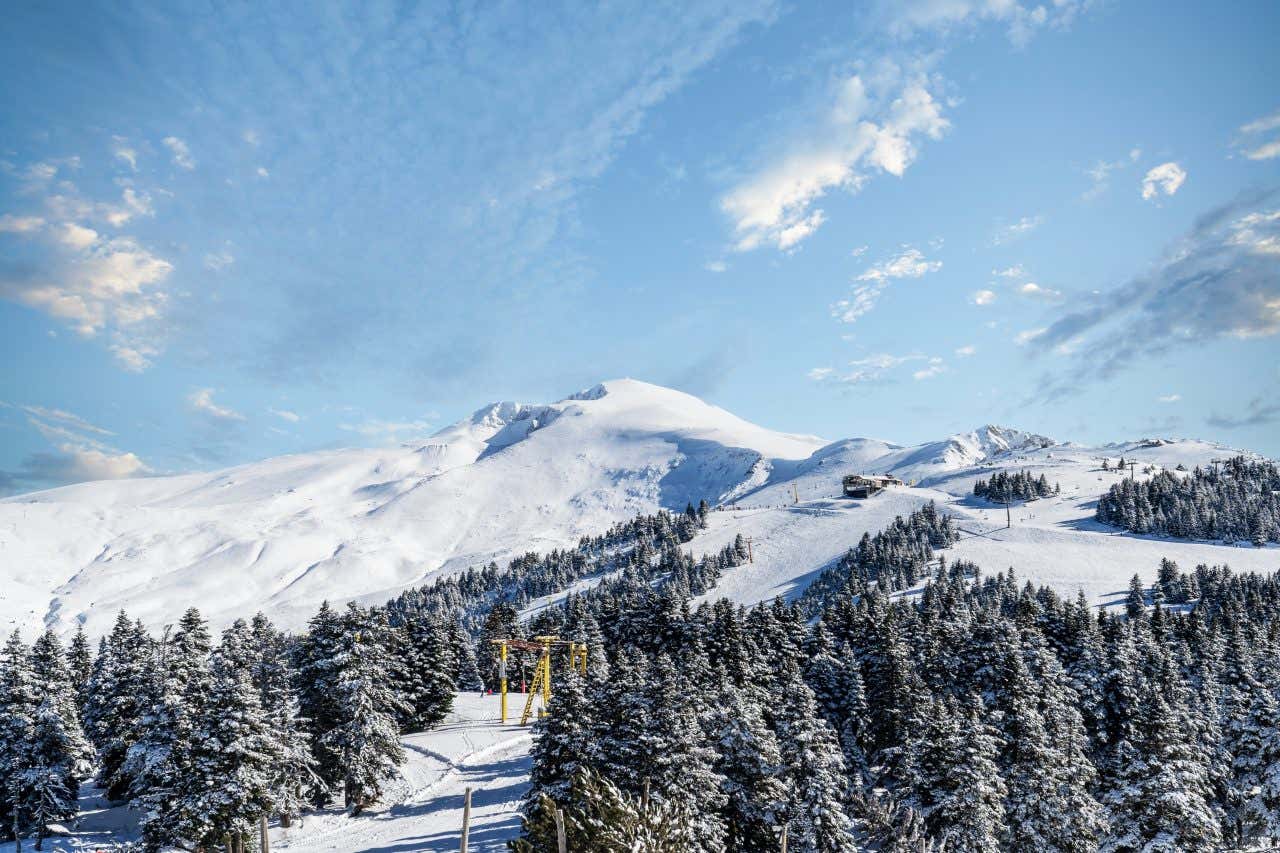A snow-covered mountain landscape with ski slopes and pine trees in the foreground. A ski lift is visible ascending the mountain, with a clear blue sky and scattered white clouds above.