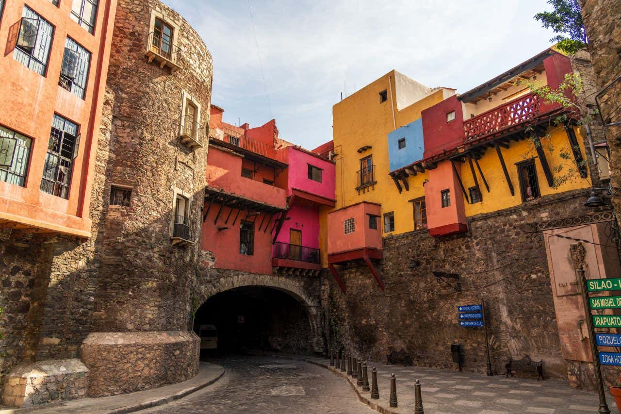 Edifício colorido construído sobre um túnel, ao lado de uma torre de pedra com um céu azul nublado ao fundo.