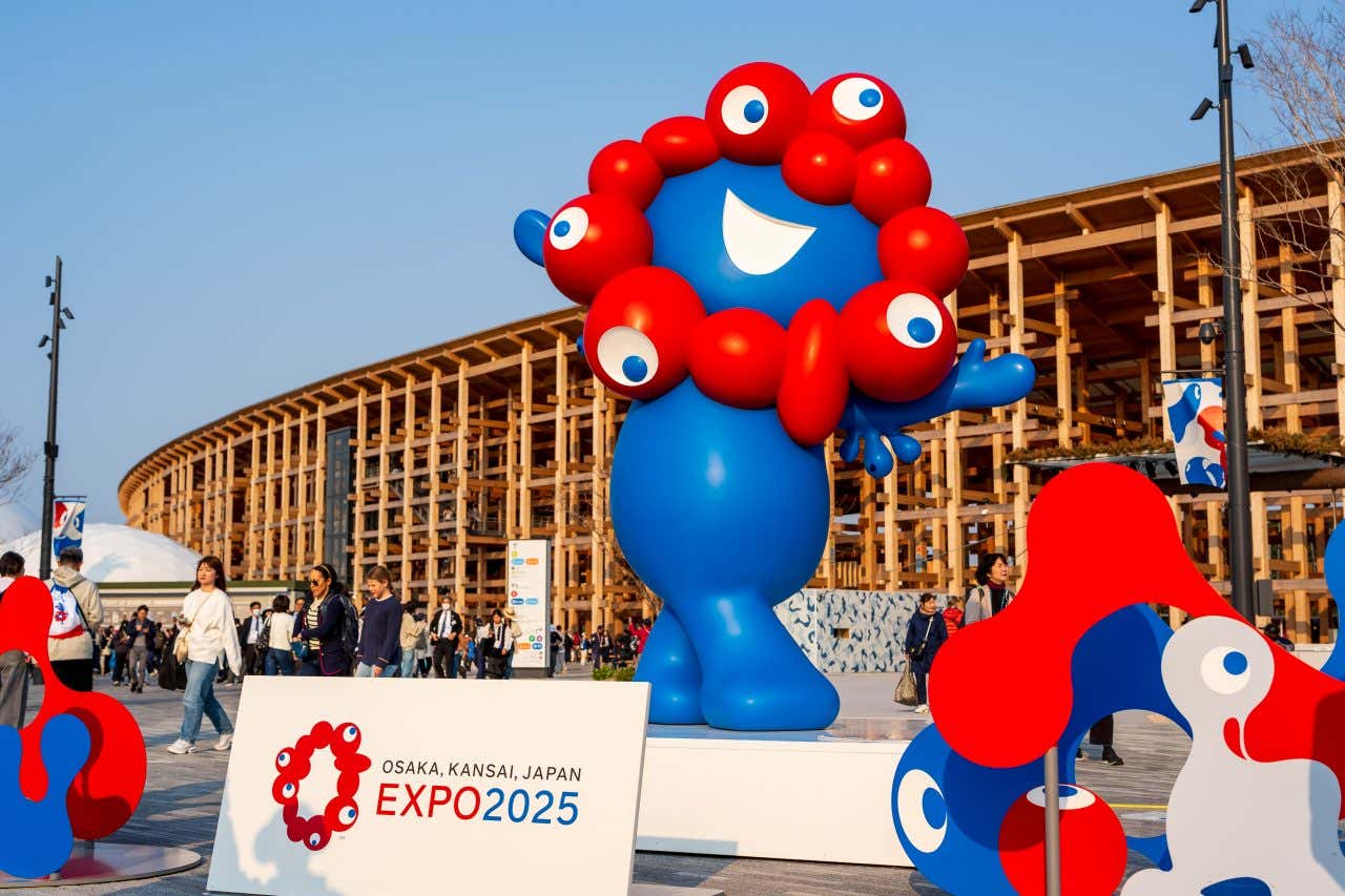 A large blue and red mascot, Myaku-Myaku, stands outdoors with an Expo 2025 sign in the foreground. People are walking in the background, and wooden structures are visible under a clear blue sky.