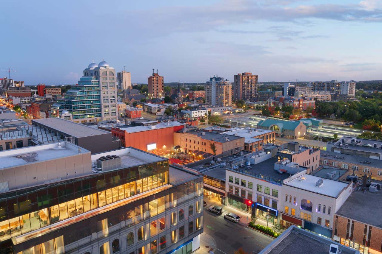 An elevated view of the city of Kitchener-Waterloo at dusk, showing a mix of modern high-rise buildings and older brick structures. Streetlights are beginning to illuminate the streets, and a residential area with green spaces is visible in the background.