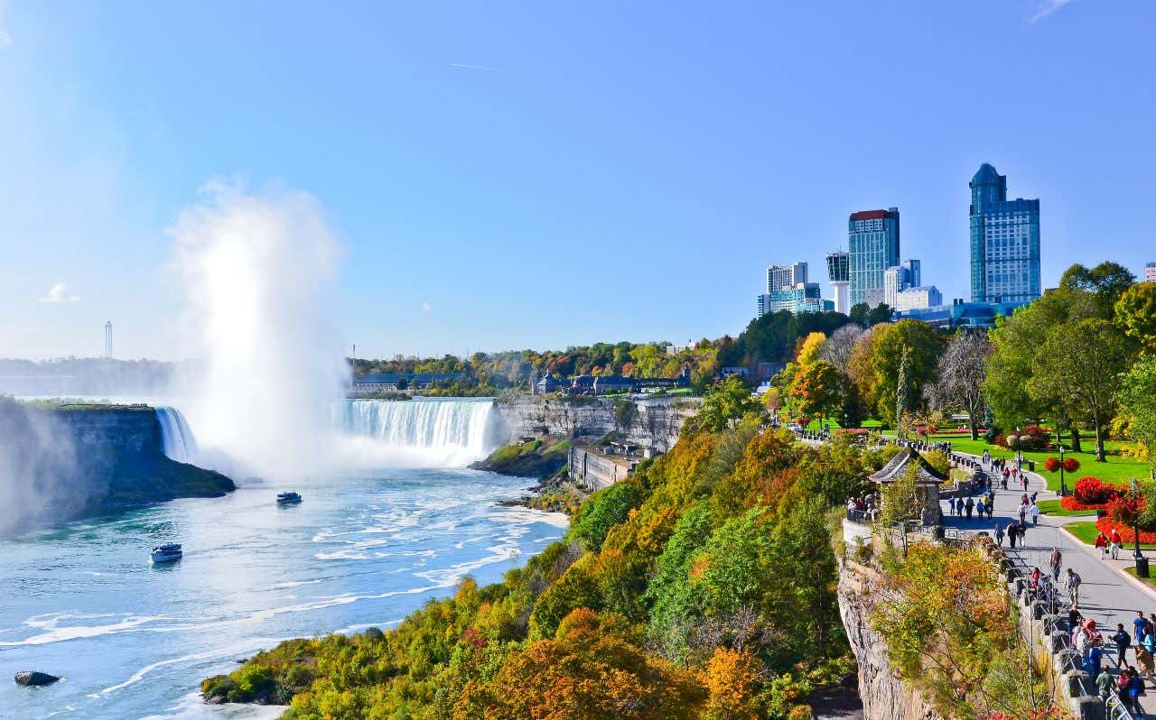 An aerial view of Niagara Falls, showing both the Horseshoe Falls and American Falls, with mist rising from the cascading water. Boats are visible on the river below. On the right, the city skyline with tall buildings and autumn foliage lines the gorge.
