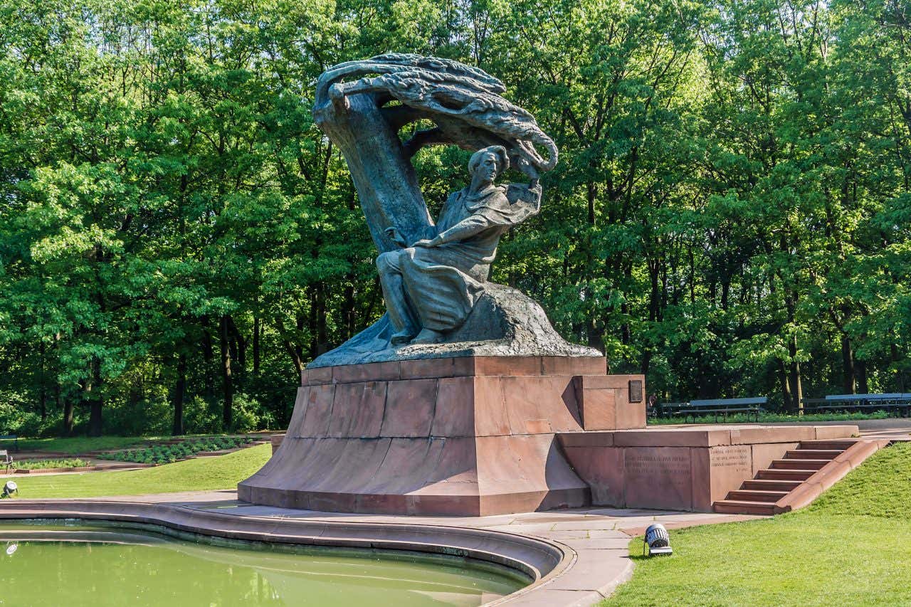 Statua di Fryderyk Chopin nel Parco Łazienki di Varsavia, raffigurante il compositore seduto sotto un salice stilizzato, i cui rami si piegano armoniosamente sopra di lui. Il monumento in stile Art Nouveau è circondato da alberi e vegetazione, in un’atmosfera tranquilla e romantica tipica del parco