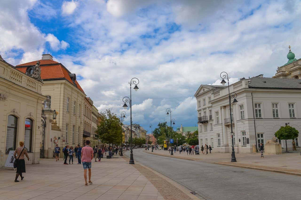 Strada Reale di Varsavia con edifici storici, lampioni decorativi ottocenteschi in ghisa nera e persone che passeggiano su un ampio marciapiede lastricato in una giornata parzialmente nuvolosa