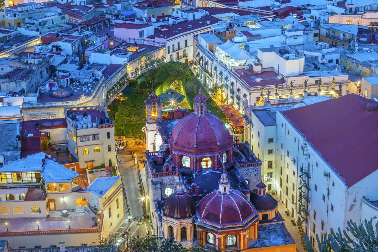 An aerial view of Guanajuato, Mexico, at dusk, showing a dense collection of buildings with various colored rooftops. Prominent in the center are two large domes and a church, with a green, arched pathway visible in the background.