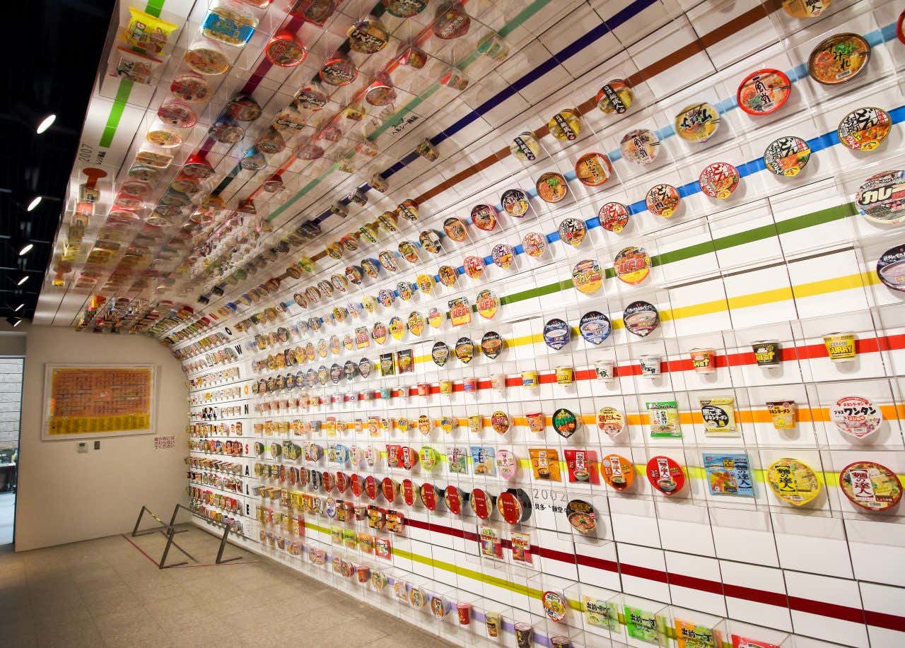 An interior view of the Momofuku Ando Instant Ramen Museum, showing a long wall display of various instant ramen packaging arranged chronologically. The ceiling also features similar displays.